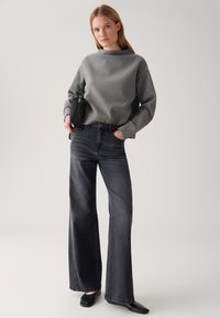 Donna in piedi con la mano destra in tasca, indossando un maglione grigio, jeans a gamba larga neri sbiaditi, scarpe nere e tenendo una piccola borsa nera.