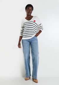 Pull blanc en V avec des rayures navy, arborant un cœur rouge brodé. Associé à un jean évasé bleu clair et des bottes à talons beiges.