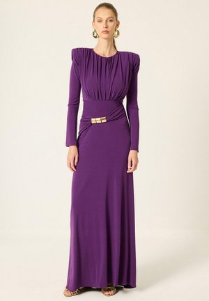Un mannequin porte une longue robe violette aux épaules froncées, une boucle dorée à la taille, de grandes boucles d’oreilles dorées et des sandales.