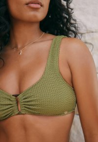 Olivegrüner Bikini-Oberteil aus strukturiertem Stoff, mit dreieckigen Cups und einem Knoten-Detail an der Vorderseite. Verfügt über dünne Träger und ein kleines goldenes Hardware-Element.