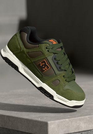 Green suede skate shoes with black accents, featuring a textured upper, padded collar, and a white rubber sole. Logo displayed in orange.