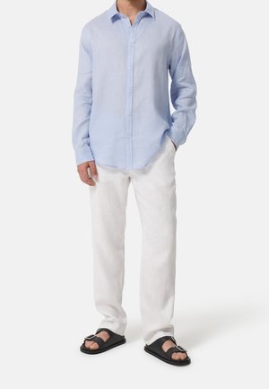 Homme portant une chemise bleu clair à manches longues boutonnée, un pantalon blanc et des sandales noires à boucle, debout avec une main dans la poche.
