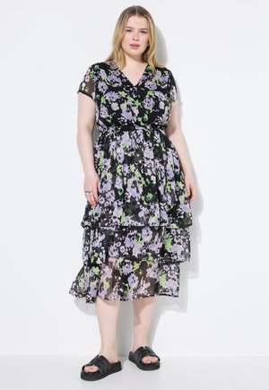 Blumenkleid in Schwarz mit lila und grünen Mustern, ausgestattet mit V-Ausschnitt, kurzen Ärmeln und geschichteten Lagen. Kombiniert mit schwarzen Sandalen.