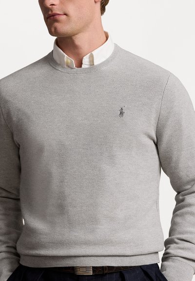 Pull en maille gris avec un col rond, un tissu texturé et un petit logo brodé sur la poitrine. Porté par-dessus une chemise blanche à col.