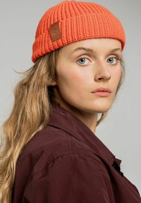 Jasnopomarańczowa dzianinowa czapka z teksturowanym, żebrowanym wzorem, z okrągłą brązową skórzaną łatą z boku.