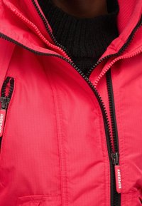 Jasnoróżowa kurtka puchowa z zamkami błyskawicznymi na piersi i kieszeniach, z teksturowanym wnętrzem i kontrastowymi czarnymi detalami zamków.