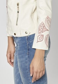 Veste en simili cuir couleur crème avec quincaillerie dorée et broderies florales rouges sur les manches, associée à un jean bleu.