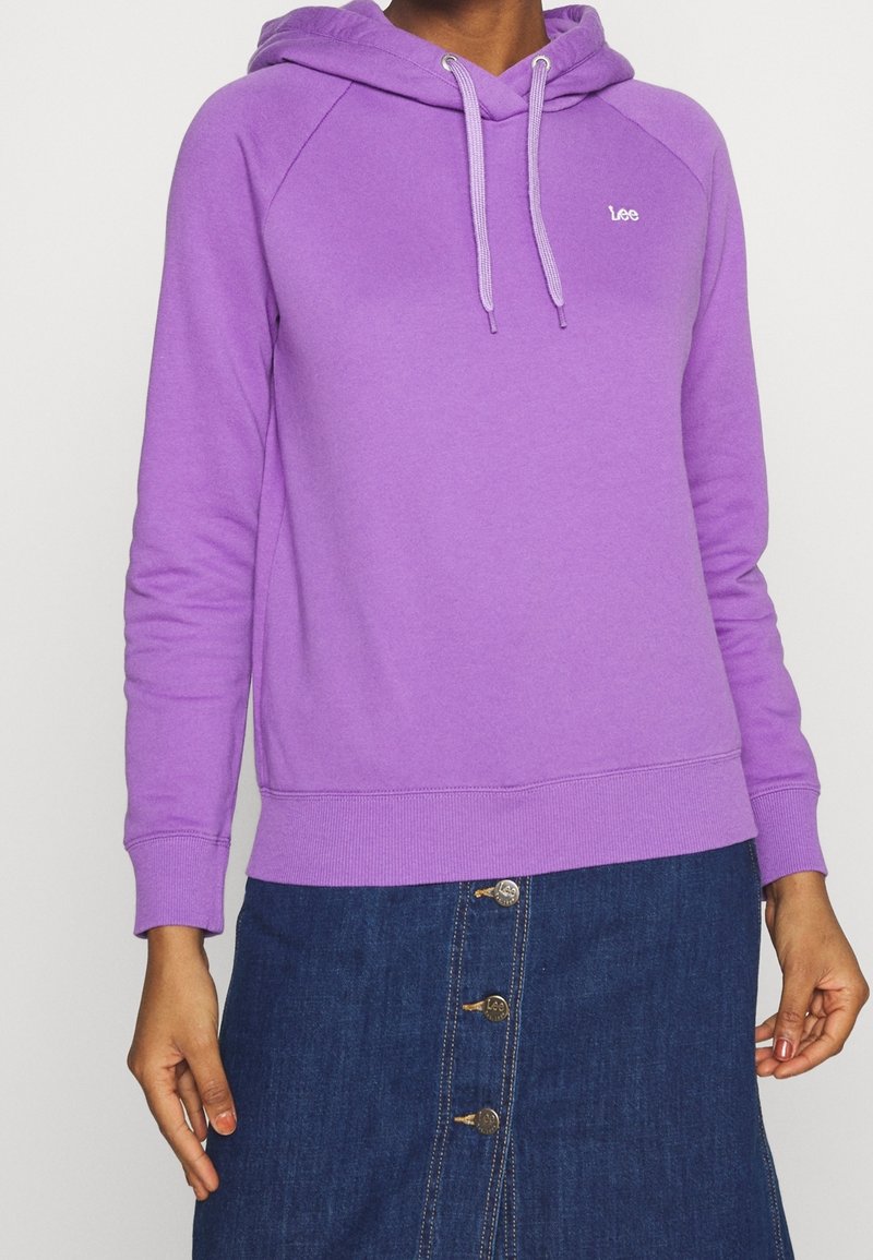 Personne portant un sweat à capuche violet avec des cordons et un petit logo blanc, associé à une jupe en jean bleue boutonnée.