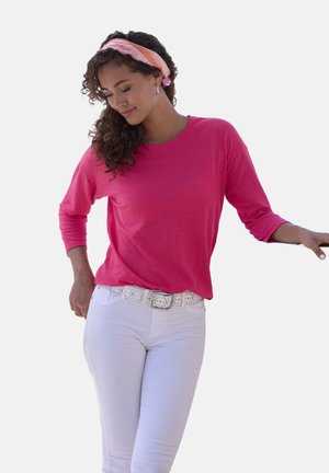 Langärmliges, leichtes rosa Oberteil mit rundem Ausschnitt, kombiniert mit weißen Hosen und einem weißen perforierten Gürtel. Model mit einem gemusterten Kopfbund.