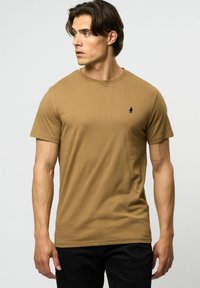 Kortermet t-skjorte med rund hals i lys brun bomull, med en liten svart logo på venstre bryst. Standard passform med en myk tekstur.