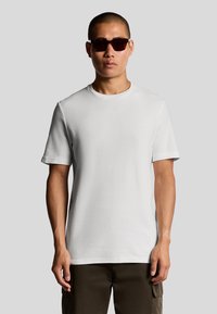 Witte korte-ik-sleeve t-shirt van gestructureerde stof, met een ronde halslijn. Model draagt een donkere zonnebril en cargobroeken.