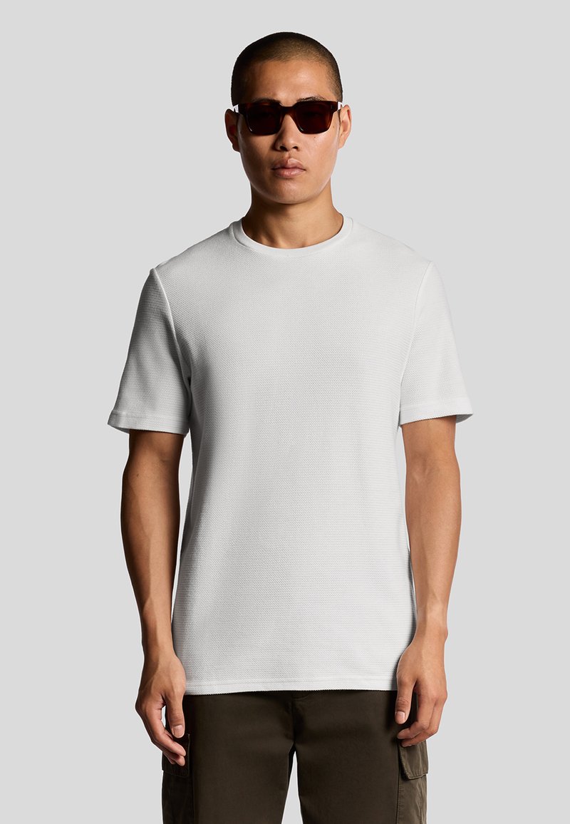 Witte korte-ik-sleeve t-shirt van gestructureerde stof, met een ronde halslijn. Model draagt een donkere zonnebril en cargobroeken.
