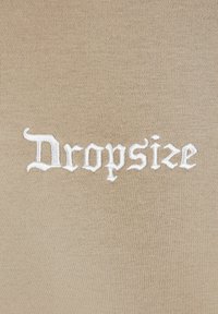 Tela beige con una textura suave que presenta un texto bordado en blanco que dice "Dropsize" en una fuente gótica estilizada.
