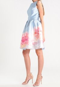 Nainen, joka on pukeutunut olkaimettomaan, vaaleansiniseen mekkoon, jossa on vaaleanpunainen kukkakuosi helmassa. Asusteena hänellä on nudenväriset, remmillä varustetut korkokengät.