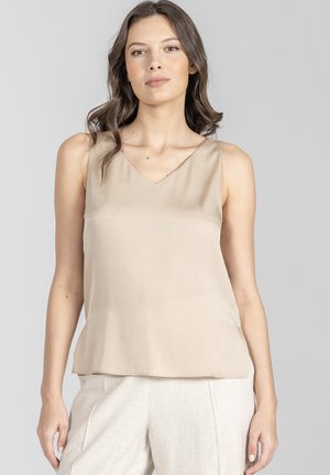 Kobieta o falujących brązowych włosach, ubrana w beżowy, bezrękawnik z dekoltem w serek oraz jasne spodnie, na jednolitym jasno-szarym tle.