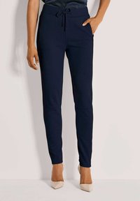 Vrouw in marineblauwe slim-fit broek met een trekkoord in de taille en zijzakken, gecombineerd met beige puntenhakken, staande tegen een effen achtergrond.