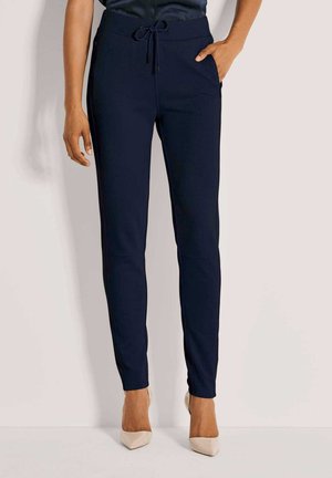 Vrouw in marineblauwe slim-fit broek met een trekkoord in de taille en zijzakken, gecombineerd met beige puntenhakken, staande tegen een effen achtergrond.