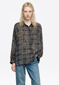 Camisa de manga larga con botones en marrón oscuro, con un diseño de cuadros azules y patrones abstractos en dorado. Corte holgado, fabricada en un tejido ligero y semi-translúcido.