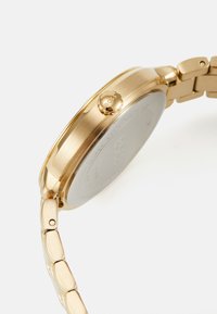 Montre-bracelet en métal doré avec un boîtier rond, finition lisse et bracelet à maillons intégrés. Présente une couronne marquée et un dos poli.