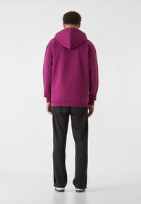 Felpa con cappuccio in magenta scuro con cappuccio regolabile, maniche lunghe e polsini a coste, abbinata a pantaloni neri a gamba affusolata.
