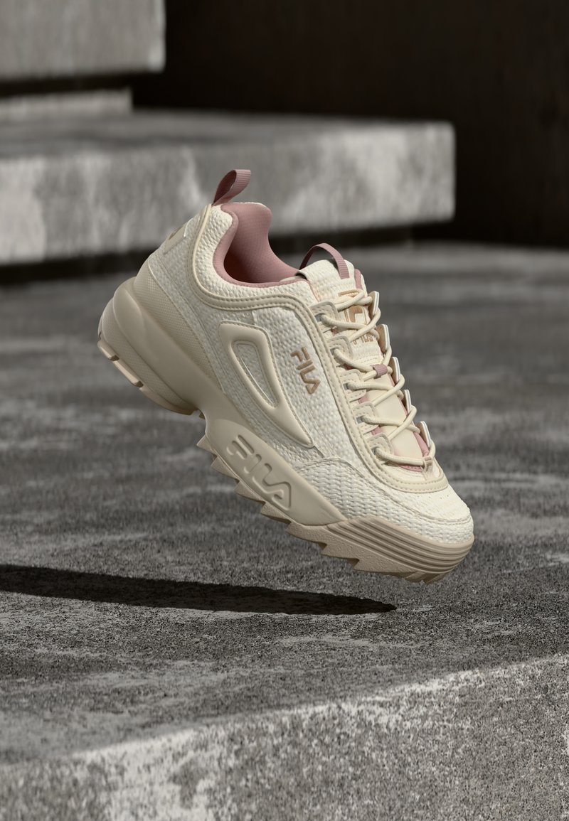 Baskets FILA crème et rose en l'air au-dessus de marches en béton, empeigne en tissu texturé, semelle épaisse avec sculptures profondes, design à lacets, et logo sur le côté.