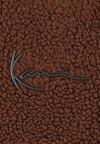 Rudas tekstūru turintis audinys su pūkuota išvaizda, kuriame įspaustas juodas stilizuotas logotipas "Kani".