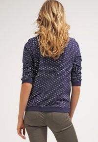 Blondynka z falującymi włosami stoi plecami, ubrana w granatową bluzkę z długim rękawem w białe kropki i szare spodnie.