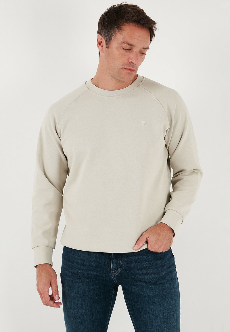 Sudadera color beige claro con mangas raglán largas y cuello redondo. Textura suave y detalle sutil del logo en el pecho. Combinada con vaqueros azules.