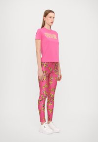 Roza majica s logotipom "Versace Jeans Couture", v kombinaciji z roza leginsami z zlatim cvetličnim vzorcem in belimi športnimi čevlji.
