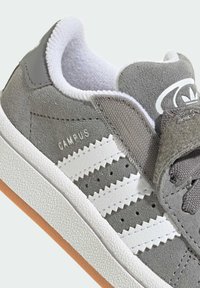 Zapatilla de ante gris con franjas de cuero blanco, collar acolchado y detalles texturizados en gris. Presenta una suela de goma blanca con una suela de goma natural.