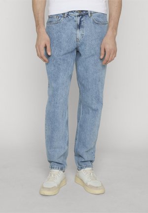 Homme portant un jean bleu clair et des baskets blanches, debout devant un fond clair uni.