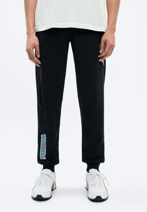 Mitad inferior de una persona con pantalones deportivos negros con el texto "PORTUGAL" en la pierna izquierda y zapatillas blancas sobre un fondo blanco.