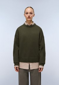 Persona che indossa un maglione di lana verde scuro sopra una camicia beige con bottoni e pantaloni grigi, in piedi contro uno sfondo chiaro e uniforme.