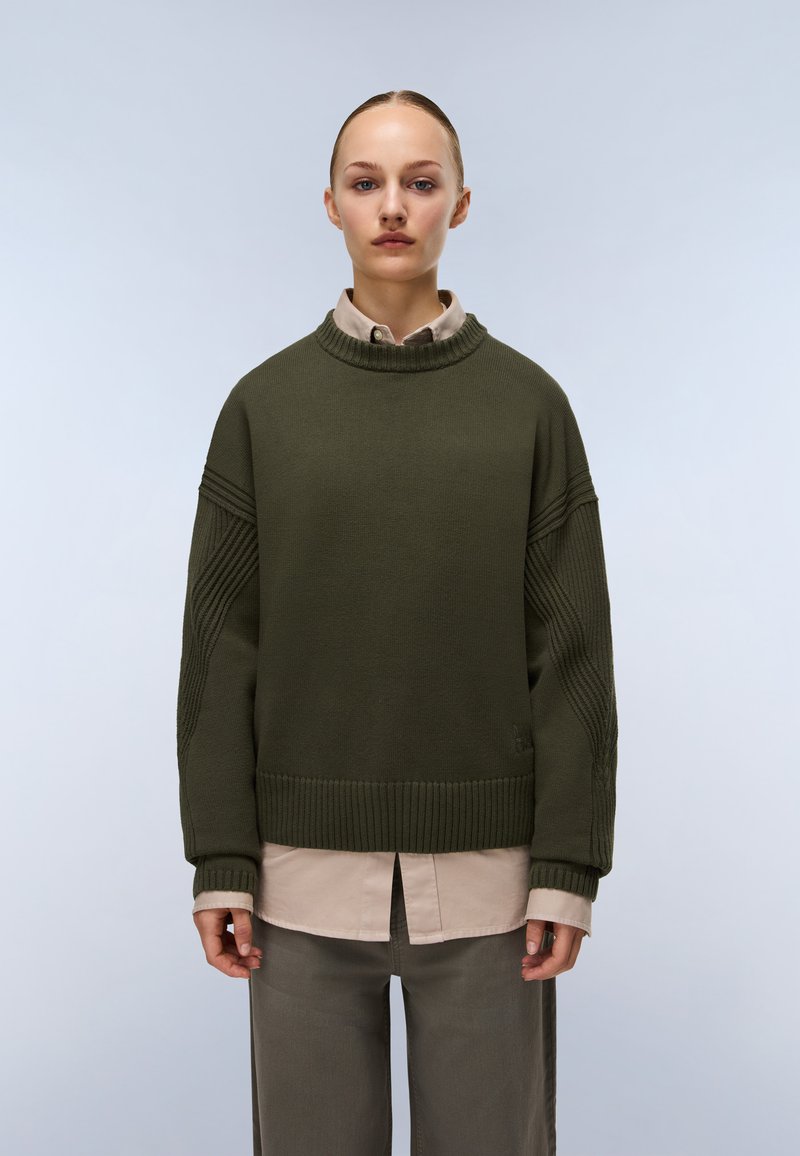 Persona che indossa un maglione di lana verde scuro sopra una camicia beige con bottoni e pantaloni grigi, in piedi contro uno sfondo chiaro e uniforme.