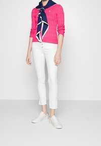 Suéter de punto trenzado rosa, jeans ajustados blancos y zapatillas blancas. Presenta una bufanda azul marino con un ribete crema alrededor del cuello.