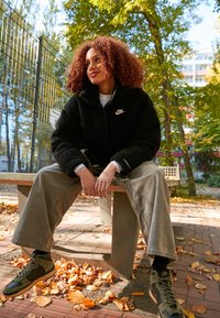 Černá fleecová bunda s logem Nike, volné světle šedé kalhoty a olivově zelené tenisky, sedící na lavičce mezi spadaným listím.