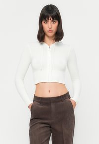 Frau mit schulterlangen, dunklen Haaren, die einen weißen, kurzen Reißverschluss-Hoodie und braune Cordhosen trägt, mit Händen in den Taschen, nach vorne gerichtet.