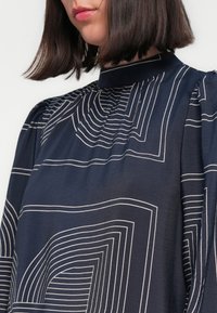 Blusa blu navy con motivi a righe bianche in diverse forme geometriche, dotata di collo alto e dettagli arricciati sulle spalle. Tessuto morbido e liscio.