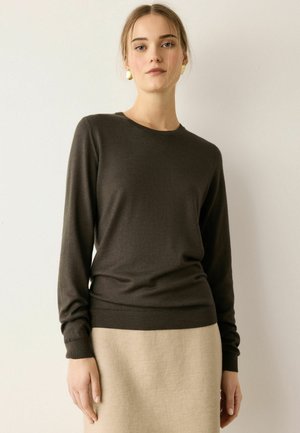 Junge Frau mit hellem Haar trägt einen dunklen Pullover mit langen Ärmeln und einen beigen Rock, sie steht vor einem schlichten hellen Hintergrund.