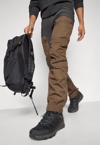 Een persoon loopt met bruine en zwarte cargobroeken, zwarte wandelschoenen en houdt een zwarte rugzak vast bij de handgreep.