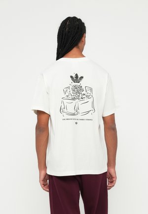 Mees punutud juustega, kandes valget t-särki, millel on restoranilaud ja Adidas logo ning tekst seljal.