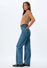 Top sin mangas de color marrón combinado con pantalones vaqueros de pierna ancha de color azul claro. El denim presenta un diseño clásico con bolsillos y un ligero desvanecimiento, terminado con zapatos negros.
