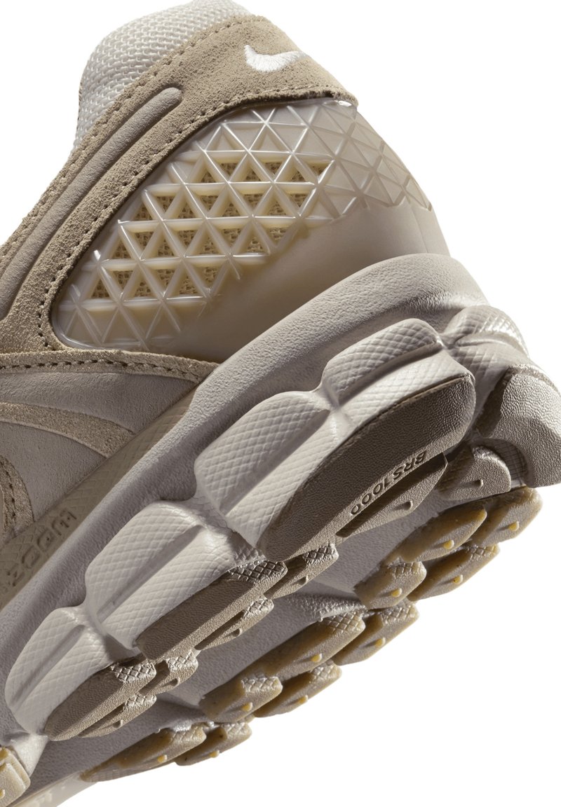 Zapatilla deportiva beige con parte superior de ante texturizado, detalles de malla y una entresuela translúcida con patrones. Suela exterior de goma con estrías en color beige claro.