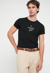 Černé bavlněné tričko s krátkým rukávem, s bílým textem "tommy girl" a hvězdným logem, spárované s béžovými kalhotami a hnědým pásem.