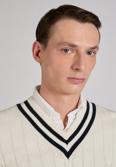 Gilet lavorato a maglia color crema con motivo a trecce, scollo a V profondo e colletto a righe blu navy, indossato sopra una camicia bianca con colletto.