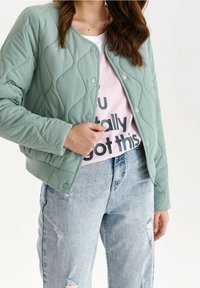Frau trägt eine hellgrüne gesteppte Jacke über einem pinken Grafik-T-Shirt und zerrissenen hellblauen Jeans, Hand passt die Vorderseite der Jacke an.