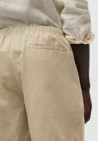 Pantaloni in cotone beige con elastico in vita e una piccola tasca a toppa sul retro, abbinati a una camicia leggera in lino. Texture morbida.