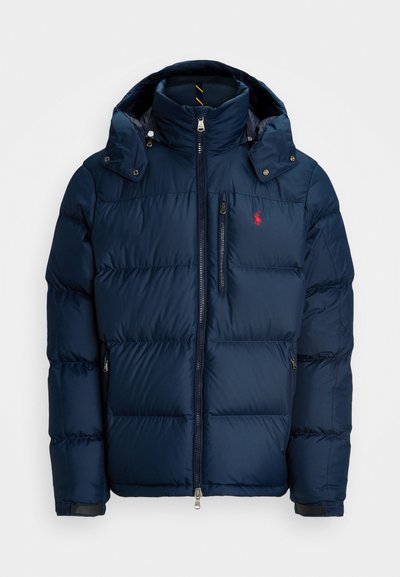 Giacca imbottita blu navy con cappuccio, dotata di chiusura con zip, tasche laterali e un piccolo logo rosso sul petto. Leggera e con finitura testurizzata.