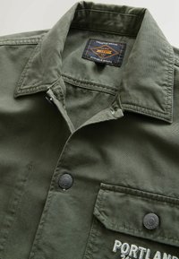 Olivgrünes Hemd mit Knopfkragen, zwei Brusttaschen, schwarzen Druckknöpfen und "PORTLAND" auf der Tasche gestickt. Strapazierfähiges Material.