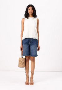 Top blanco de punto con un patrón texturizado, combinado con shorts de denim hasta la rodilla y sandalias de tiras color beige. Los accesorios incluyen un bolso de mimbre.